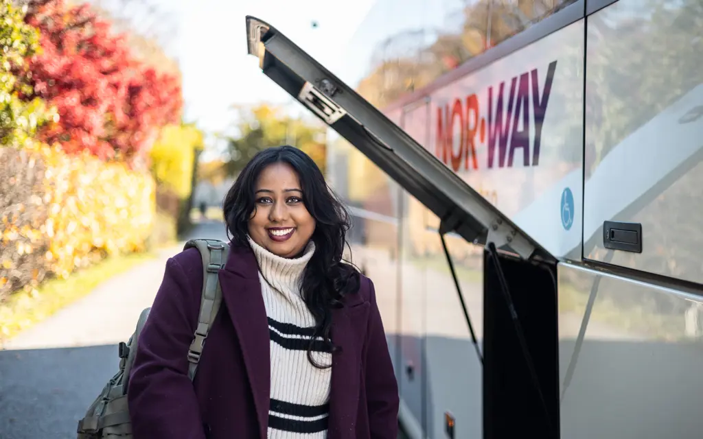 Jente står foran NOR-WAY-buss i høstfarger