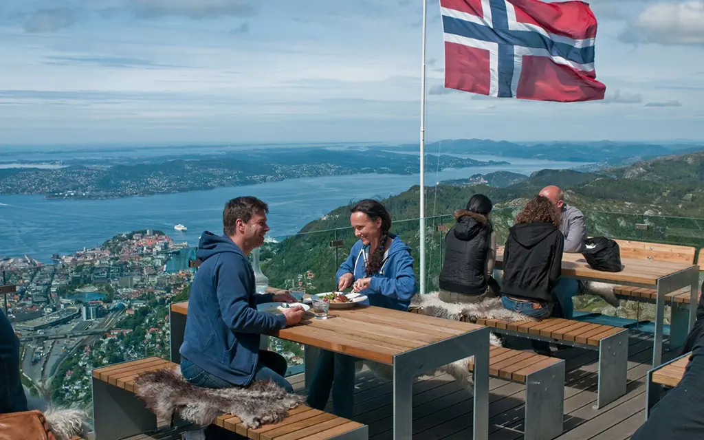 Mennesker spiser mat ved bord på utkikkspunkt over Bergen by