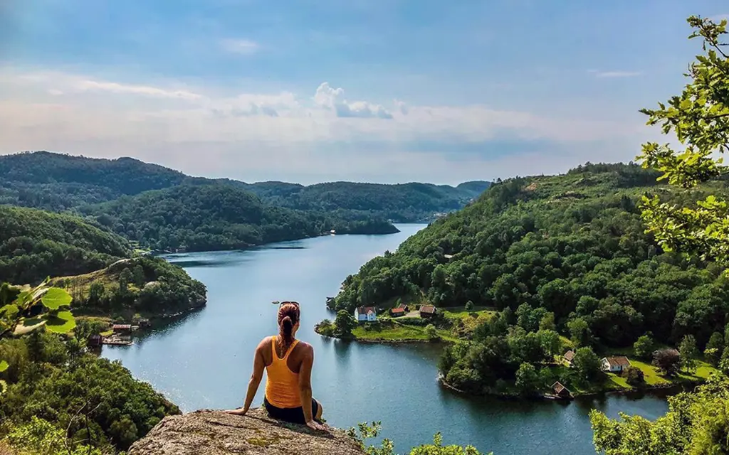 The woman looks out over a landscape with a lake, forest, and hills.