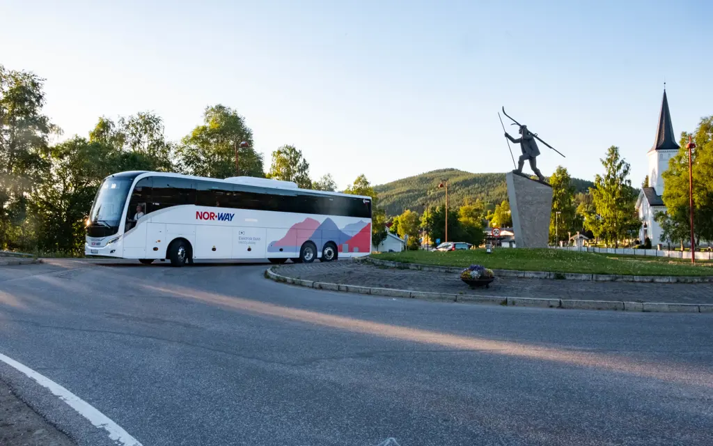 Elbuss Trysilekspressen i rundkjøring i Trysil med kirke i bakgrunnen og Trysil-Knut statue i forgrunnen