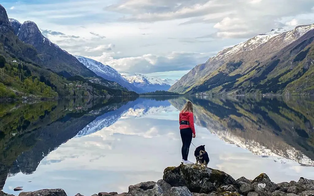 Kvinne og hund foran stille vann og fjellandskap