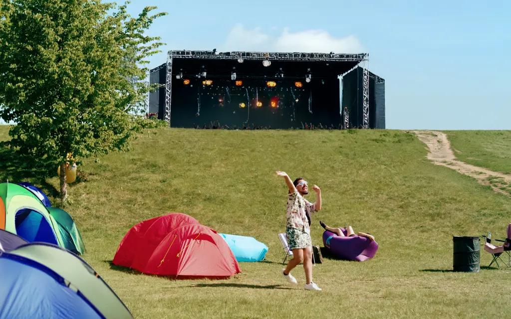 Dansende mann på en grønn gresslette mellom telt med konsertscene i bakgrunnen på festival